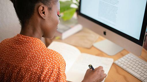 Looking over a students shoulder as they write in a notebook while sitting in front of a computer